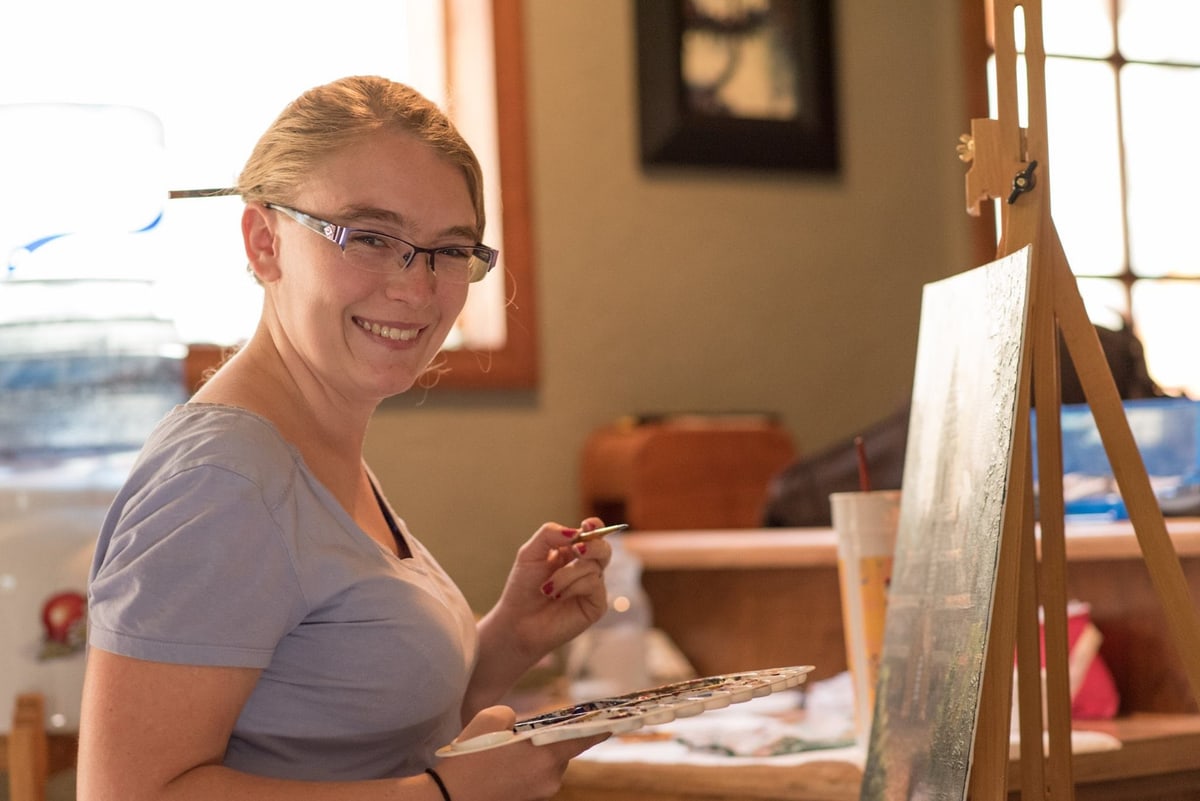 Stephanie Fowler in her studio, smiling at the camera with a paintbrush and palette